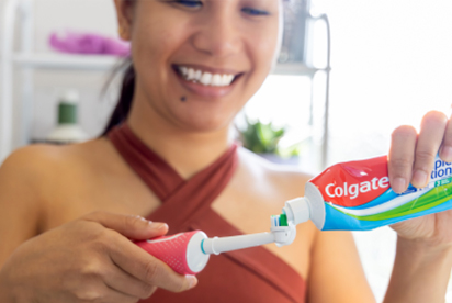 Una mujer sonríe mientras pone pasta de dientes Colgate en un cepillo de dientes en un baño.